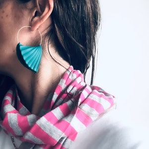 Pode incluir: Uma mulher usando um cachecol listrado rosa e cinza com um brinco em forma de leque azul turquesa. O brinco está preso a um aro de ouro.