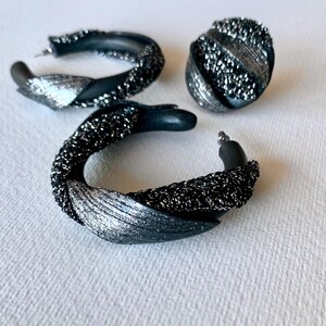 Pode incluir: Um conjunto de três peças de joalharia: dois brincos de argola e um brinco de pressão. Os brincos são pretos com detalhes de glitter prateado. Os brincos de argola têm um design torcido, e o brinco de pressão tem formato redondo.