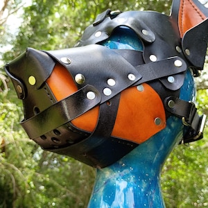 May include: A black and orange leather animal-style mask featuring pointed ears, a snout-like protrusion, and silver-toned metal rivets. The mask is constructed from layered leather straps and is displayed on a blue, glittery mannequin head.