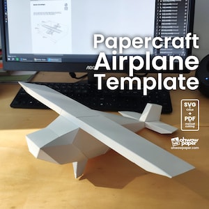 Puede incluir: Un modelo de avión de papel blanco sobre una mesa de madera. El avión está ensamblado y tiene un diseño detallado. La imagen también muestra una pantalla de computadora con una plantilla de avión de papel. El texto en la pantalla dice "Plantilla de avión de papel". El texto en la mesa dice "SVG para Cricut + PDF corte manual ohwowpaper.com".