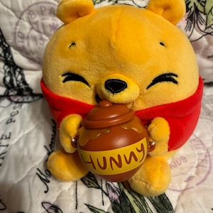 May include: A plush yellow bear toy with a round head and body, wearing a red shirt. The bear has embroidered black eyes and a black nose, and is holding a brown plastic honey pot with a yellow label that has the word HUNNY printed on it.