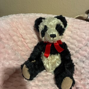 May include: A plush panda bear with black and white fur, a red bow, and tan paws. The stuffed animal is sitting on a pink, textured surface. The panda's face has black markings around the eyes and ears.