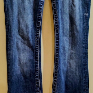 Op de afbeelding: Een paar donkerblauwe spijkerbroeken met een bootcut-poot. De jeans heeft een decoratief stikseldetail langs de naden en een klein versiering op de achterzak.