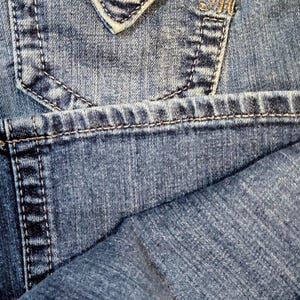 Op de afbeelding: Blauwe denim jeans met een decoratief zakje met gouden en zwarte pailletten en een zilveren knoop. De jeans hebben een Miss Me merklabel.