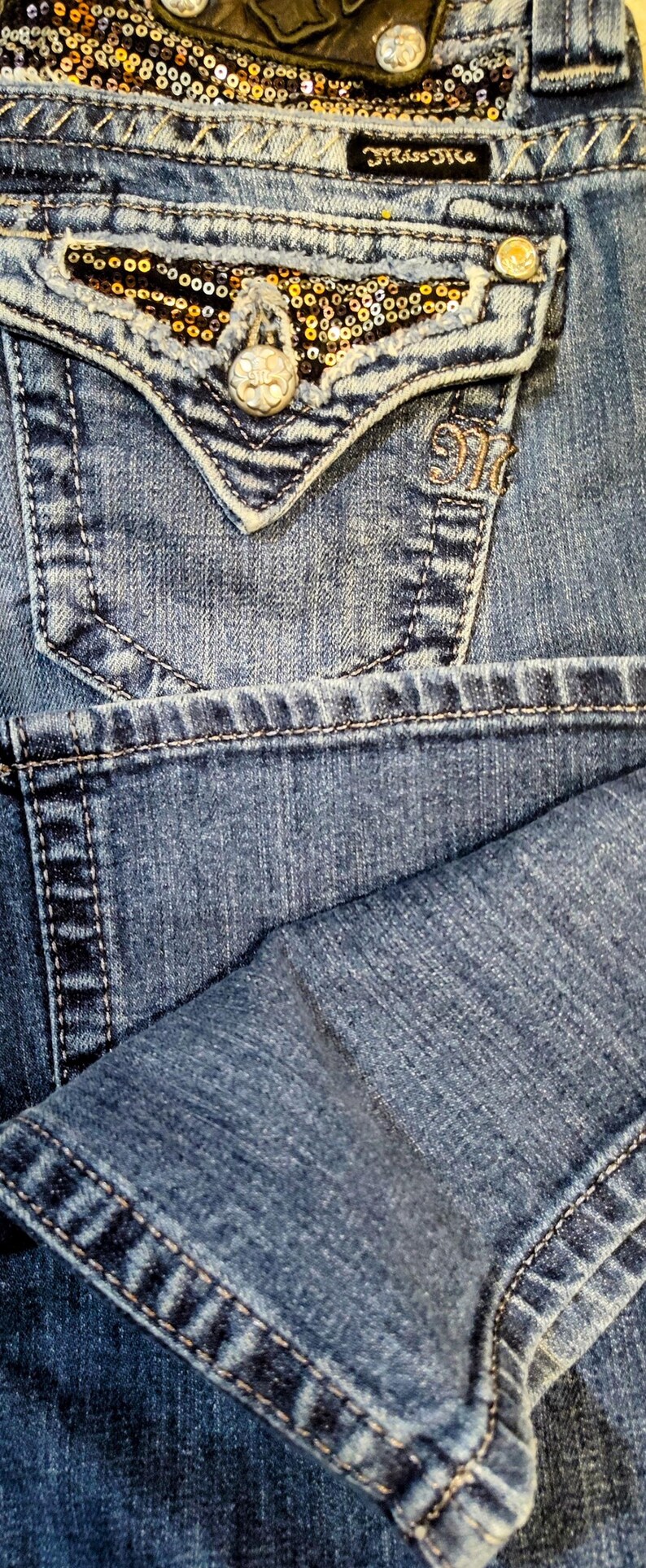 Op de afbeelding: Blauwe denim jeans met een decoratief zakje met gouden en zwarte pailletten en een zilveren knoop. De jeans hebben een Miss Me merklabel.