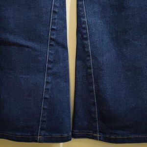 May include: Close-up of the lower portion of two dark blue denim jean legs. The jeans have a slight flare at the bottom and are stitched with a contrasting yellow thread. The seams are visible.