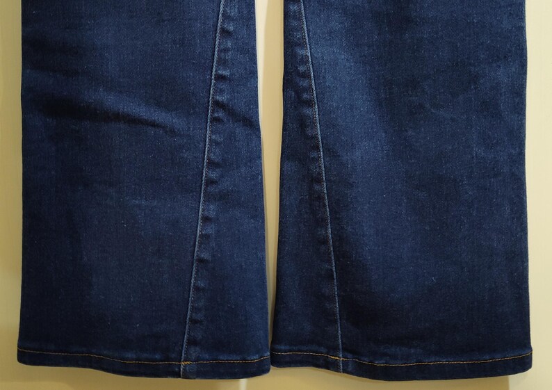 May include: Close-up of the lower portion of two dark blue denim jean legs. The jeans have a slight flare at the bottom and are stitched with a contrasting yellow thread. The seams are visible.