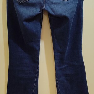 May include: Dark blue denim jeans with a bootcut leg. The back pockets feature a gold-colored stitched design. The waistband and pockets are accented with gold stitching. The jeans are hanging on a metal hanger.