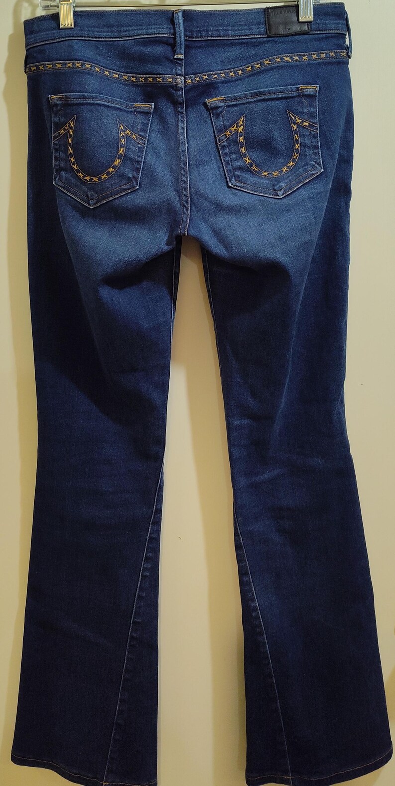 May include: Dark blue denim jeans with a bootcut leg. The back pockets feature a gold-colored stitched design. The waistband and pockets are accented with gold stitching. The jeans are hanging on a metal hanger.