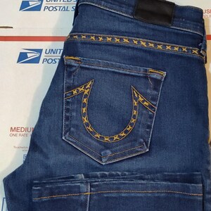 May include: Dark blue denim jeans with yellow stitching details. The back pocket features a distinctive horseshoe design with yellow stitching. The jeans have a black leather patch and are folded. The jeans are on top of a cardboard box.