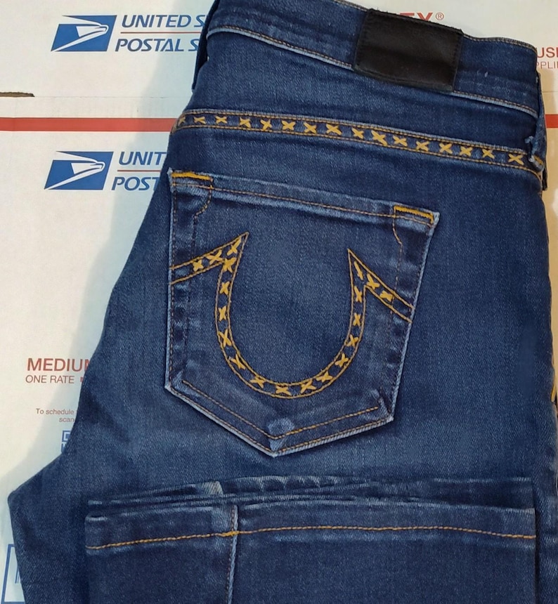 May include: Dark blue denim jeans with yellow stitching details. The back pocket features a distinctive horseshoe design with yellow stitching. The jeans have a black leather patch and are folded. The jeans are on top of a cardboard box.