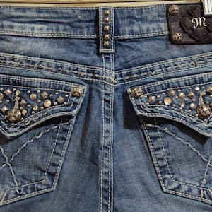 May include: Close-up of the back of blue denim jeans. The jeans feature decorative stitching, embellished back pockets with silver studs and rhinestones, and a leather patch with the initials "MS".
