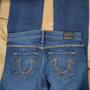 May include: Dark blue denim jeans with a flared leg. The back pockets feature a yellow embroidered design. The waistband has a black leather patch. The jeans are laid flat on a gold-colored surface.