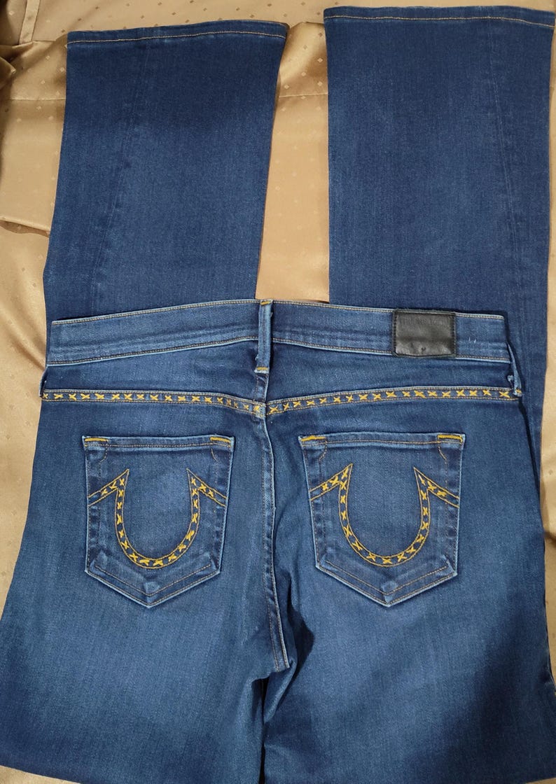 May include: Dark blue denim jeans with a flared leg. The back pockets feature a yellow embroidered design. The waistband has a black leather patch. The jeans are laid flat on a gold-colored surface.
