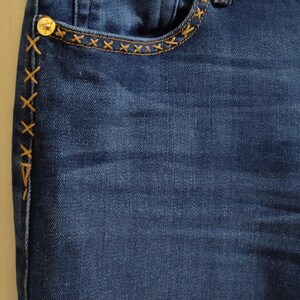May include: Close-up of dark blue denim jeans. The jeans feature a small pocket with a decorative stitch pattern and a gold button. The side seam has a decorative stitch pattern. The jeans have a classic five-pocket design.