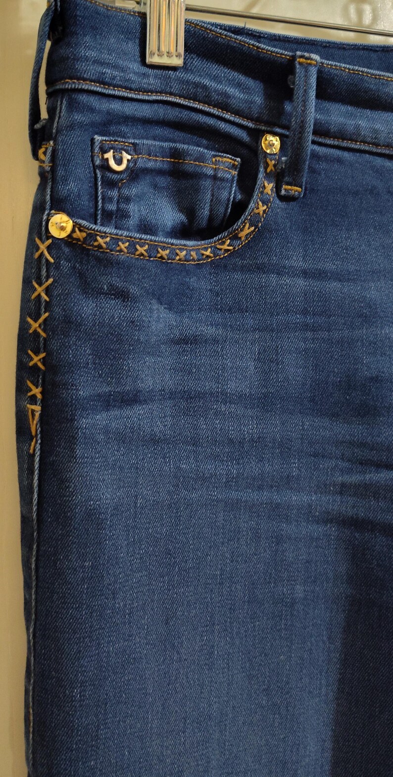 May include: Close-up of dark blue denim jeans. The jeans feature a small pocket with a decorative stitch pattern and a gold button. The side seam has a decorative stitch pattern. The jeans have a classic five-pocket design.