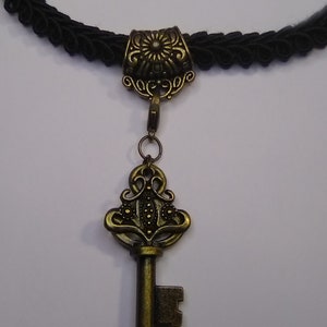 May include: Black choker necklace with a bronze-colored key pendant. The key has an ornate design, and the necklace features a woven black cord. The pendant is attached to the choker with a small clasp.
