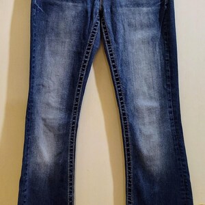 Op de afbeelding: Een paar blauwe denim jeans met een bootcut-poot. De jeans heeft een lichte wassing en is distressed met een klein gat op het rechterbeen. De jeans heeft een zilveren knoop en een rits.
