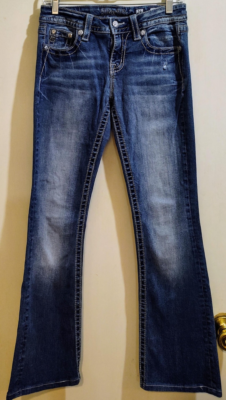 Op de afbeelding: Een paar blauwe denim jeans met een bootcut-poot. De jeans heeft een lichte wassing en is distressed met een klein gat op het rechterbeen. De jeans heeft een zilveren knoop en een rits.