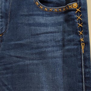 May include: Close-up of blue denim jeans with decorative gold stitching. The jeans feature gold hardware, including buttons and rivets. The pocket edges and side seams are accented with cross-stitch details.