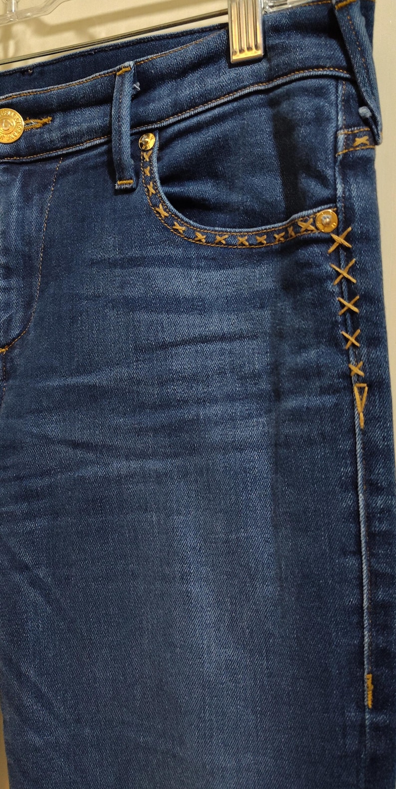 May include: Close-up of blue denim jeans with decorative gold stitching. The jeans feature gold hardware, including buttons and rivets. The pocket edges and side seams are accented with cross-stitch details.