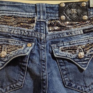 Op de afbeelding: Blauwe denim jeans met paillettendetails op de achterzakken en de tailleband. De achterzakken hebben een kenmerkende V-vorm en de tailleband heeft een zwart leren patch met de tekst "M".