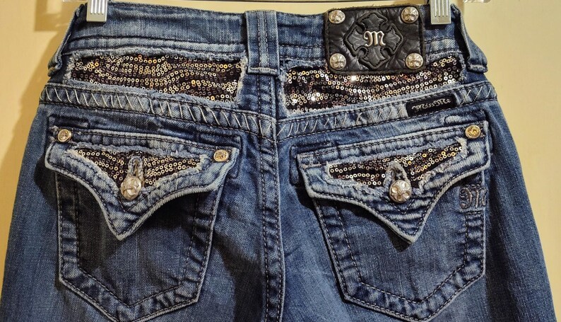 Op de afbeelding: Blauwe denim jeans met paillettendetails op de achterzakken en de tailleband. De achterzakken hebben een kenmerkende V-vorm en de tailleband heeft een zwart leren patch met de tekst "M".