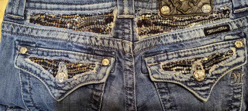 Op de afbeelding: Blauwe denim jeans met zwarte en gouden paillettendetails op de achterzakken en de tailleband. De jeans hebben een klein leren patch met het woord "Miss Me" op de achterkant van de tailleband.