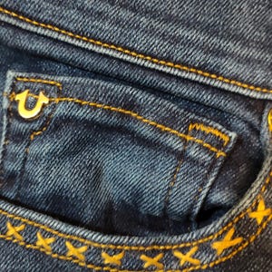 May include: Close-up of blue denim jeans with gold stitching. The jeans have a small pocket with a gold logo and a label that reads "BECCA" and "Mid Rise Bootcut Mi-taille Bottillon."