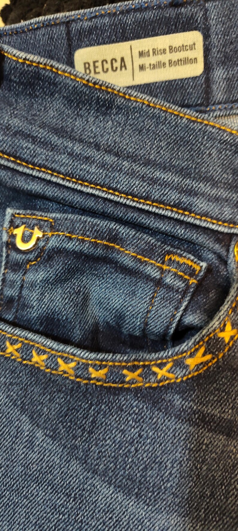 May include: Close-up of blue denim jeans with gold stitching. The jeans have a small pocket with a gold logo and a label that reads "BECCA" and "Mid Rise Bootcut Mi-taille Bottillon."