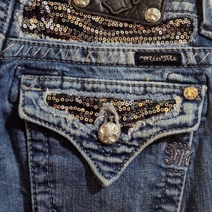 Op de afbeelding: Blauwe denim jeans met een zilveren knoop en een zwart leren patch met een zilveren M erop. De jeans hebben een zak met een gouden en zwarte pailletten patch.