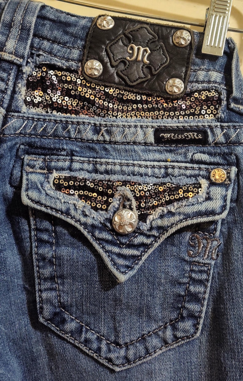 Op de afbeelding: Blauwe denim jeans met een zilveren knoop en een zwart leren patch met een zilveren M erop. De jeans hebben een zak met een gouden en zwarte pailletten patch.