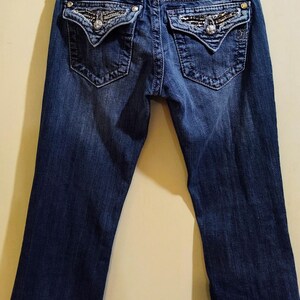 Op de afbeelding: Een donkerblauwe spijkerbroek met een paillettenband en versierde achterzakken. De broek heeft een bootcut-stijl en heeft het "JR"-logo op de achterkant.