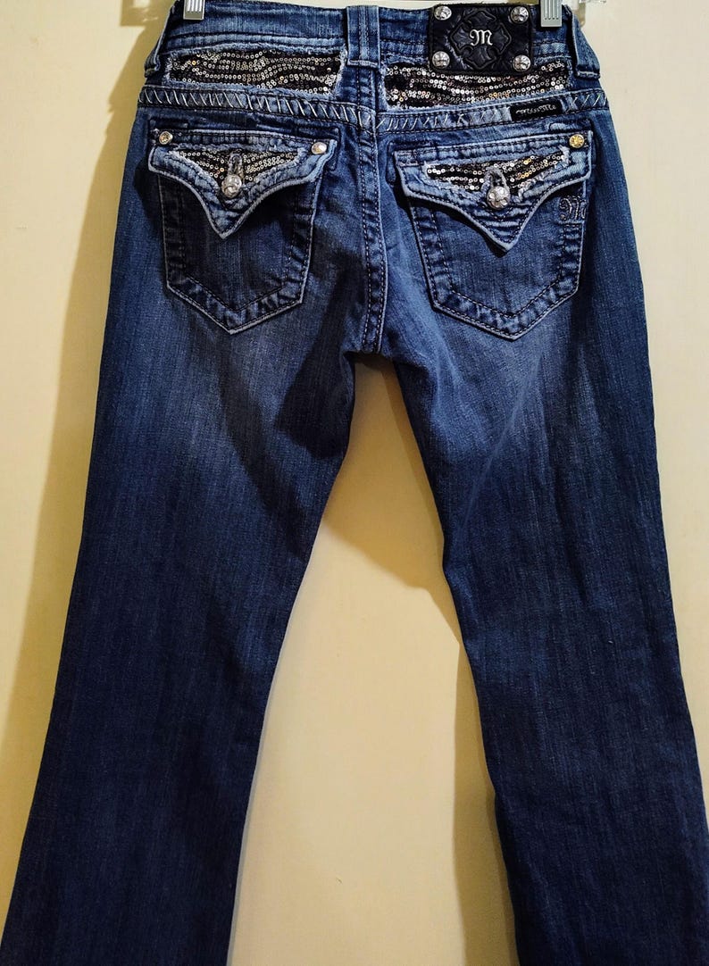Op de afbeelding: Een donkerblauwe spijkerbroek met een paillettenband en versierde achterzakken. De broek heeft een bootcut-stijl en heeft het "JR"-logo op de achterkant.