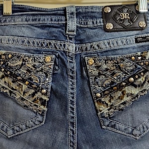 May include: Blue denim jeans with decorative stitching and embellished back pockets. The pockets feature a mix of patterns, studs, and metallic accents. A leather patch with the brand logo is visible at the waistband.