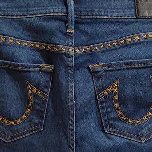 May include: Close-up of the back of blue denim jeans. The jeans feature two back pockets with a horseshoe design and decorative stitching. The waistband and pockets have gold-colored stitching. A black leather patch is visible.