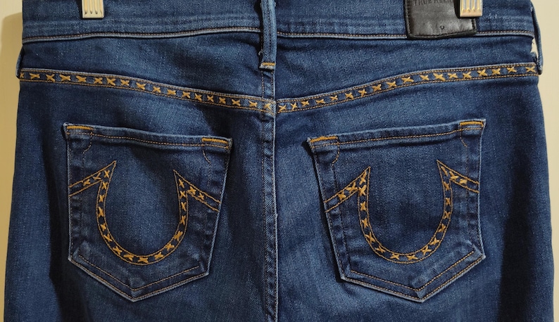 May include: Close-up of the back of blue denim jeans. The jeans feature two back pockets with a horseshoe design and decorative stitching. The waistband and pockets have gold-colored stitching. A black leather patch is visible.