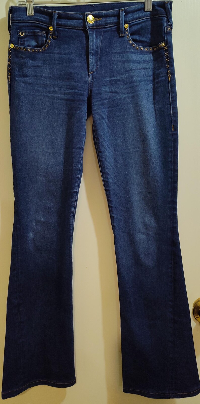 May include: Dark wash denim jeans with a bootcut leg. The jeans have gold-colored hardware, including rivets and a button. Decorative stitching in gold thread accents the pockets and side seams.