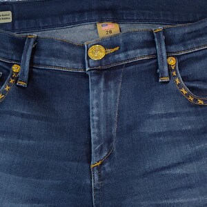 May include: Dark blue denim jeans with gold-colored hardware and decorative stitching. The jeans feature a button closure, front and back pockets, and a size 28 tag. The stitching is in a star pattern.