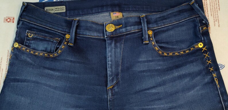 May include: Dark blue denim jeans with gold-colored hardware and decorative stitching. The jeans feature a button closure, front and back pockets, and a size 28 tag. The stitching is in a star pattern.