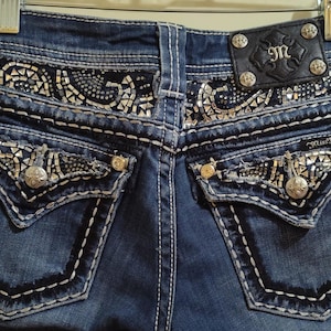 Può includere: Primo piano del retro di jeans blu con cuciture e abbellimenti decorativi. Presenta borchie argentate, motivi intricati e una toppa in pelle con il logo del marchio. I jeans sono appesi a una gruccia di metallo.
