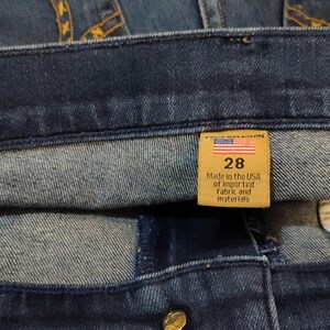 May include: Close-up of dark blue denim jeans with a mid-rise bootcut. A tan tag reads "BECCA" and "Mid Rise Bootcut". Another tag shows "28" and "Made in the USA of imported fabric and materials".