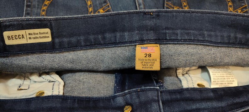 May include: Close-up of dark blue denim jeans with a mid-rise bootcut. A tan tag reads "BECCA" and "Mid Rise Bootcut". Another tag shows "28" and "Made in the USA of imported fabric and materials".