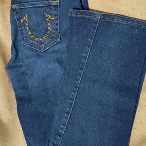 May include: Dark blue denim jeans with a bootcut leg. The back pocket features a yellow stitched design. Yellow stitching accents the back yoke and along the seams. The jeans have a classic five-pocket design and a leather patch.