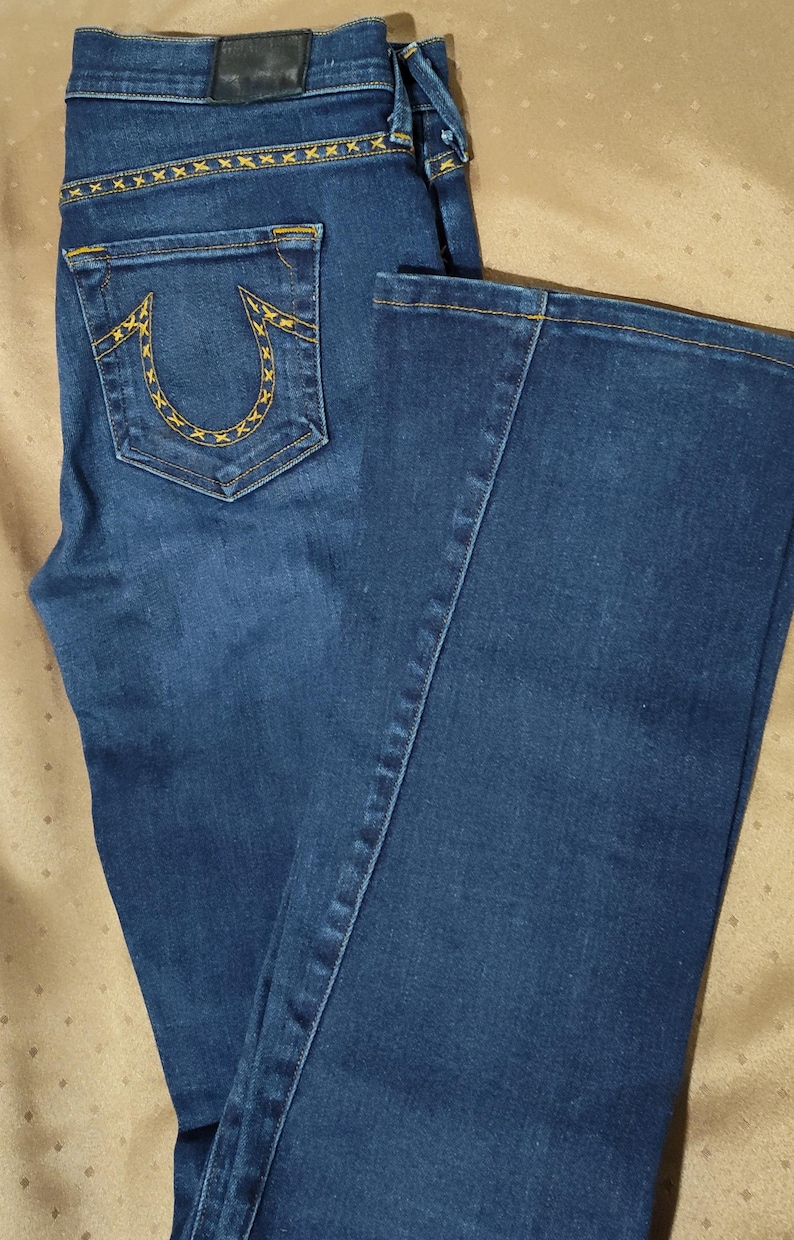 May include: Dark blue denim jeans with a bootcut leg. The back pocket features a yellow stitched design. Yellow stitching accents the back yoke and along the seams. The jeans have a classic five-pocket design and a leather patch.