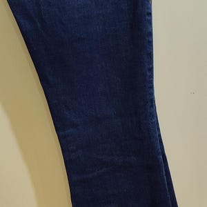 May include: Dark blue denim jeans with a flared leg. The jeans have a decorative stitch design on the back pockets and waistband. The jeans are hanging on a metal hanger.