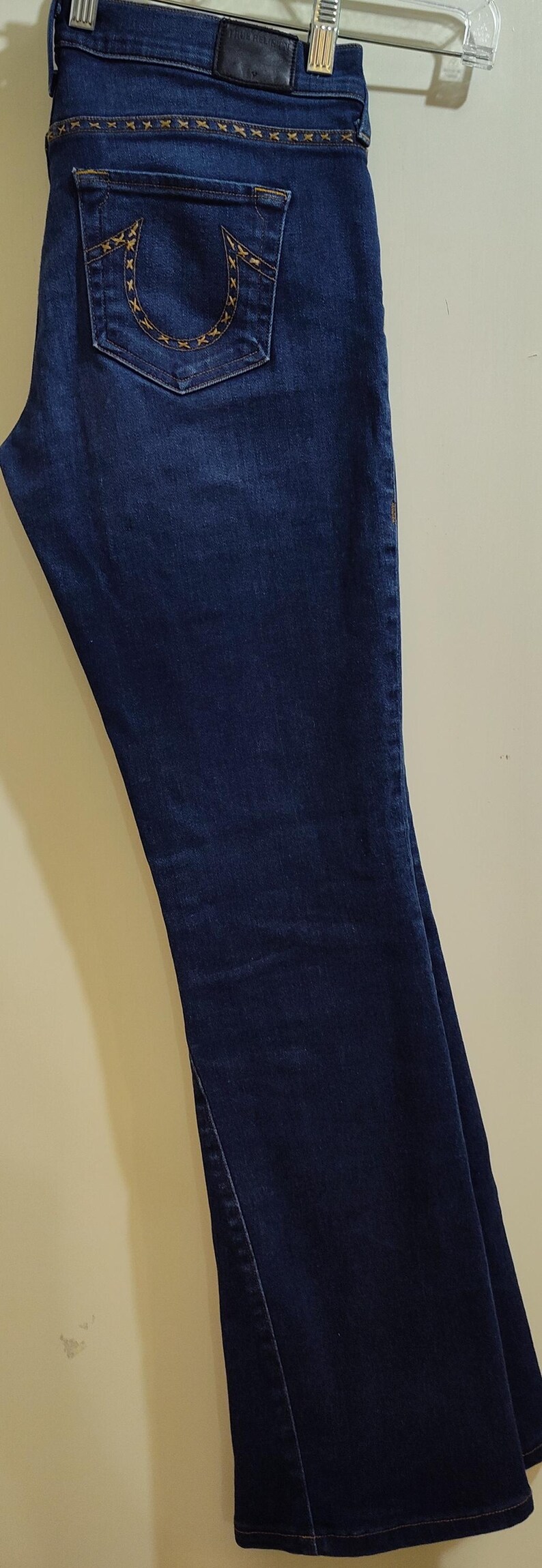May include: Dark blue denim jeans with a flared leg. The jeans have a decorative stitch design on the back pockets and waistband. The jeans are hanging on a metal hanger.