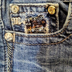 Op de afbeelding: Close-up van een blauwe denimzak met een zilveren M-logo, een gouden paillettenpatch en twee goudkleurige knopen.