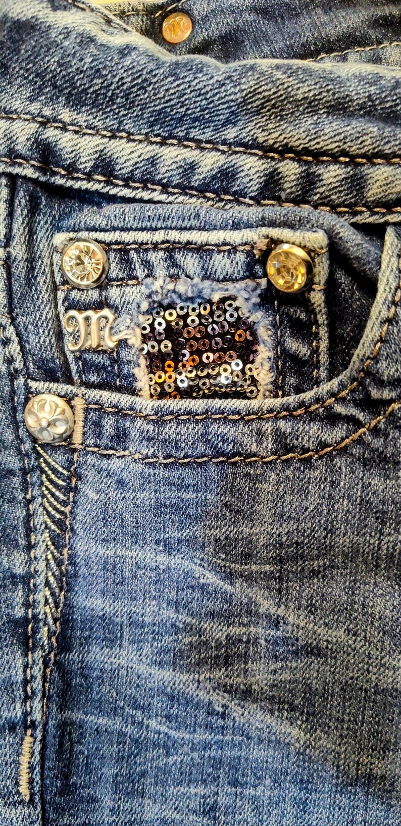 Op de afbeelding: Close-up van een blauwe denimzak met een zilveren M-logo, een gouden paillettenpatch en twee goudkleurige knopen.