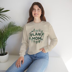Puede incluir: Sudadera beige con el texto "PLANT MOM" en verde, rodeada por una corona de ilustraciones de plantas verdes. La parte superior de manga larga es usada por una persona con vaqueros azules.